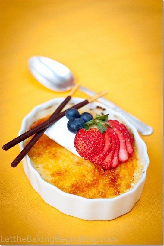 Creme brulee in a white decorative bowl topped with strawberries and blueberries with two chocolate sticks with a spoon.