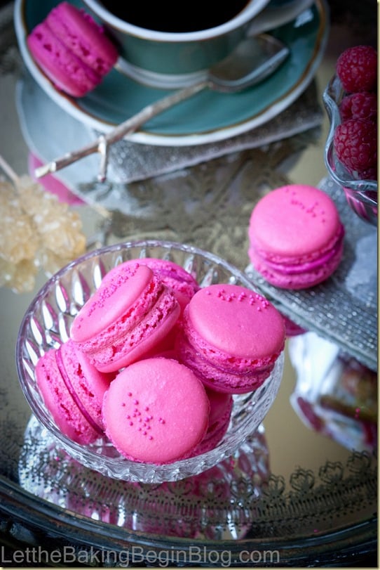 French macarons with raspberries in a glass bowl next to a cup of black coffee.