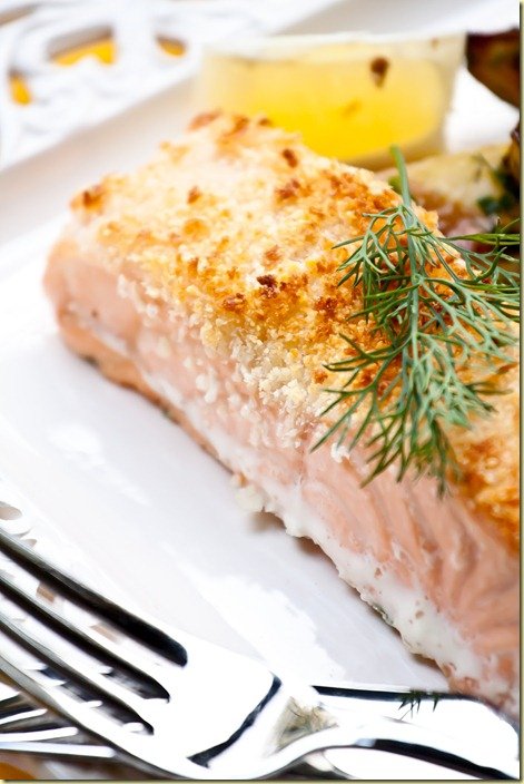 Side view of parmesan crusted salmon topped with fresh greens on a white plate with fresh lemons.