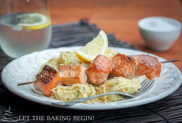  Side view of a bowl of spaghetti noodles and oven grilled salmon kabobs and a fresh lemon on a white decorative bowl.