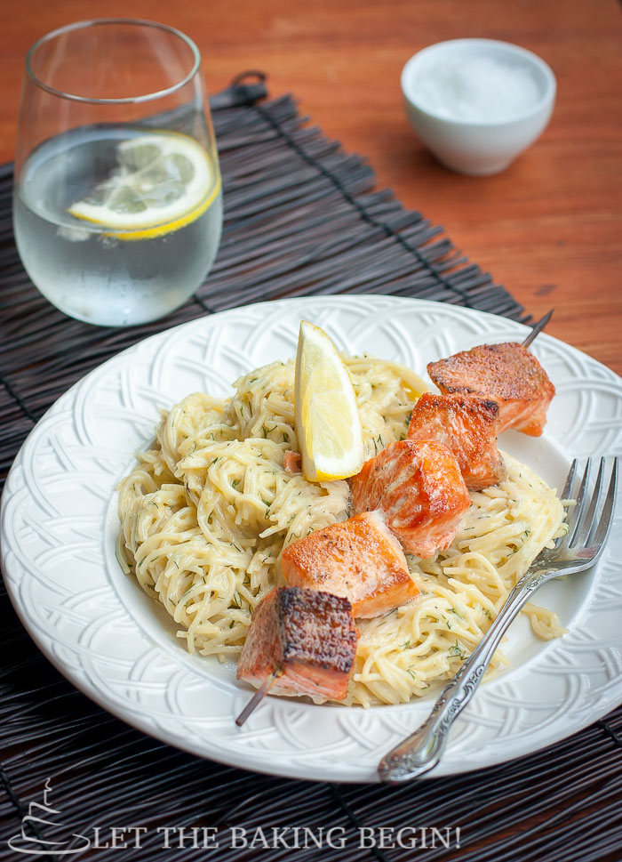 Oven grilled salmon kabobs sided with spaghetti noodles and a fresh lemon in a white decorative bowl. 