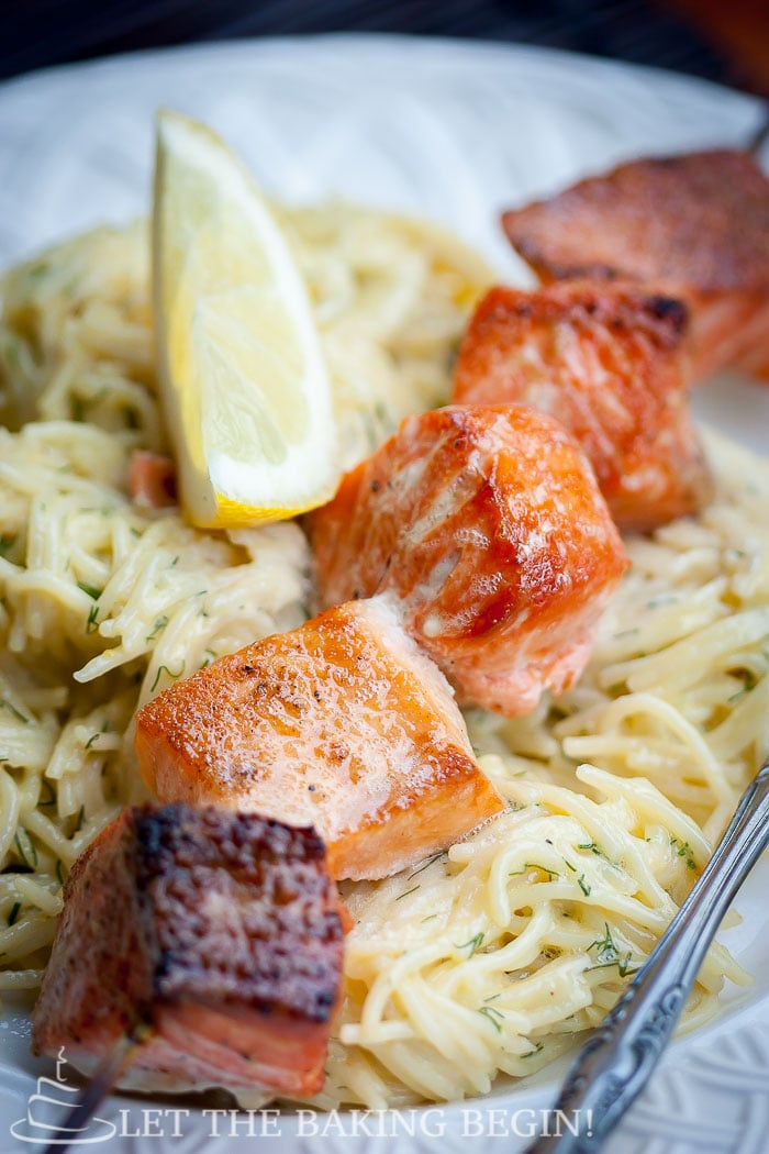 Side view of oven grilled salmon kabobs on top of spaghetti noodles and a fresh lemon in a white decorative bowl. 