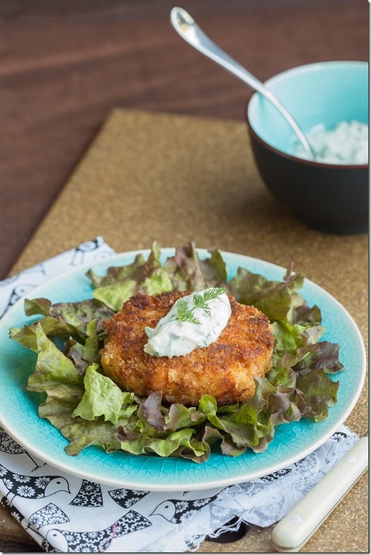 Salmon cake on top of plate of romaine topped with tzatziki sauce and fresh greens on a decorative plate.