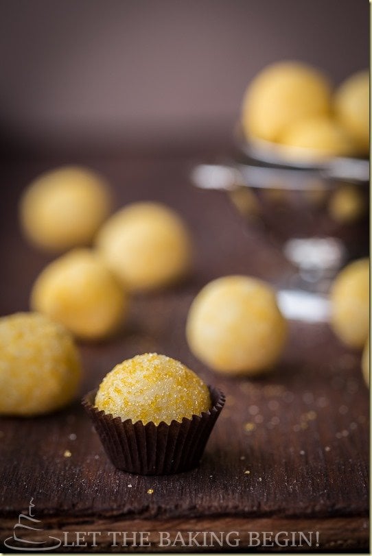 Lemon truffles in a brown liner on a brown table with scattered lemon truffles in the background.
