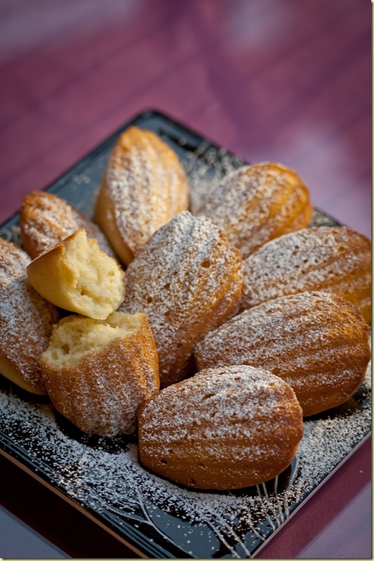 Lemon madeleines with one  broken in half on a black decorative plate topped with powdered sugar.