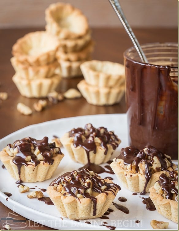  Tartlets with walnuts and topped with chocolate on a white tray and empty tartlets with jar of melted chocolate.