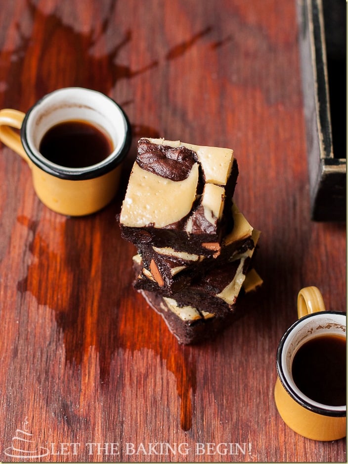 Cream cheese brownies stacked on a table with spilled coffee and two coffee mugs.