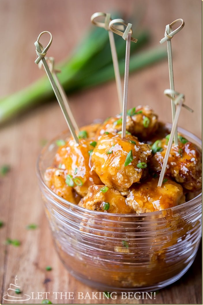 Chinese orange chicken in a glass bowl with skewers in the chicken topped with fresh greens and sesame seeds.