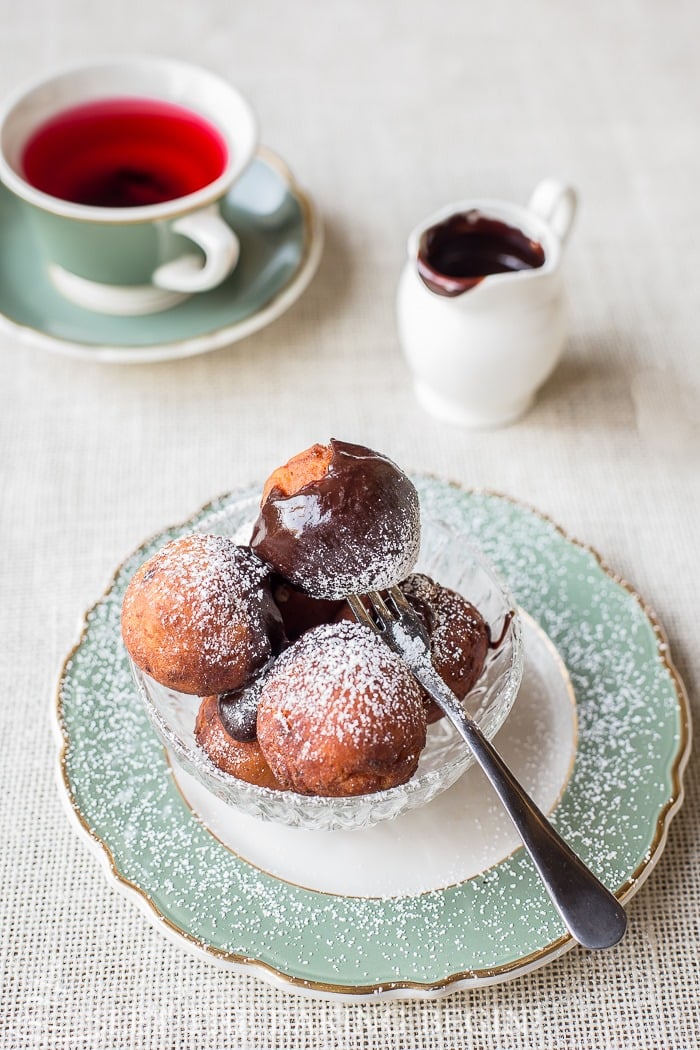 Top view of donuts topped with chocolate ganache and powdered sugar in a glass bowl on a green and white decorative plate, with a cup of tea, and chocolate ganache in a white pouring cup.