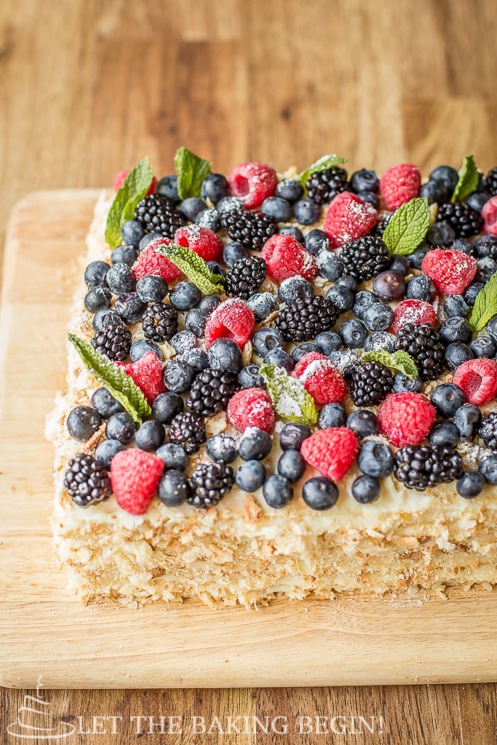 Cake topped with blueberries, raspberries, blackberries, blueberries, mint, and powdered sugar on a wooden cutting board. 