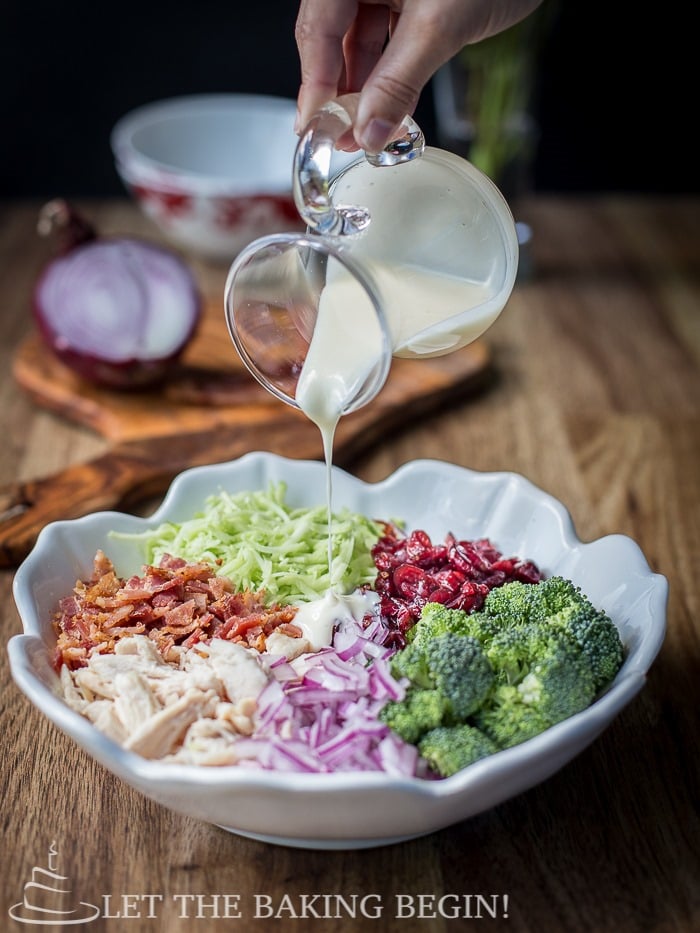 Dressing being poured on broccoli salad in a white decorative bowl on a wooden table. 