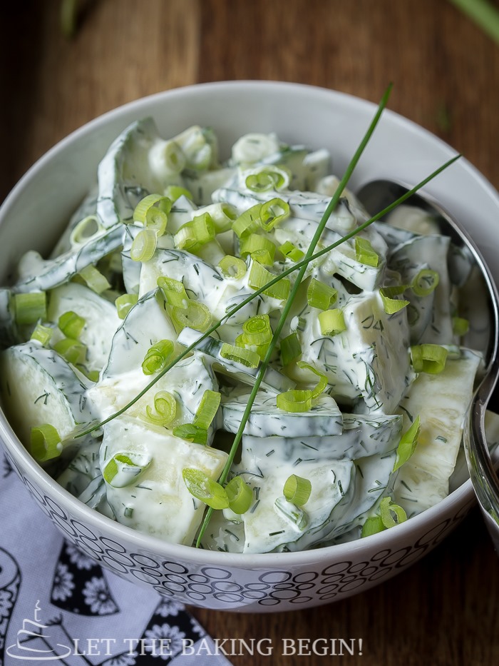  Top view of cucumber dill salad topped with fresh greens in a white decorative bowl on a wooden table.