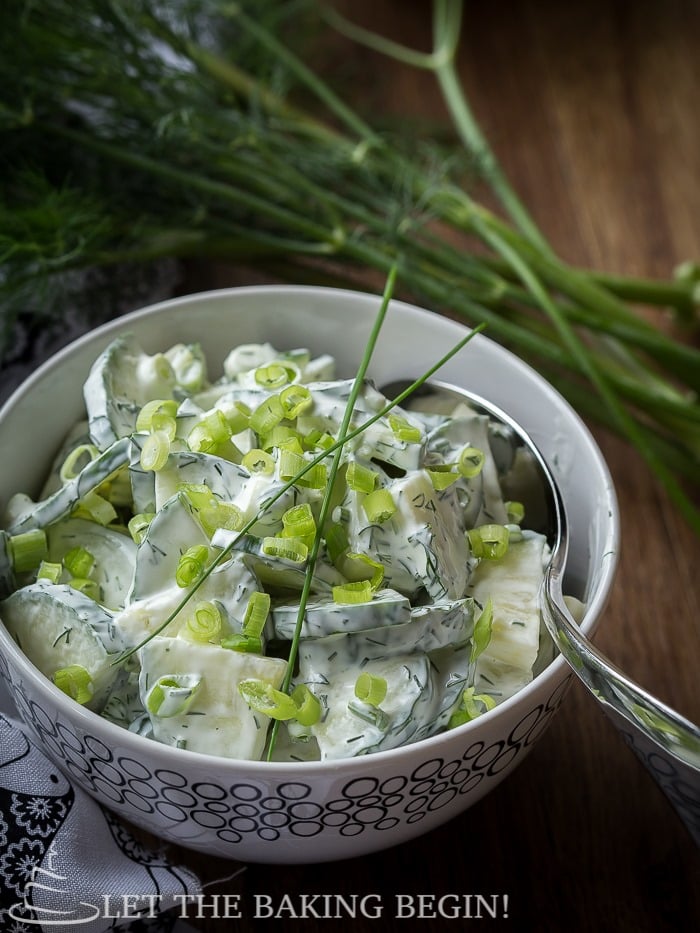 Cucumber dill salad topped with fresh greens in a white decorative bowl on a wooden table.