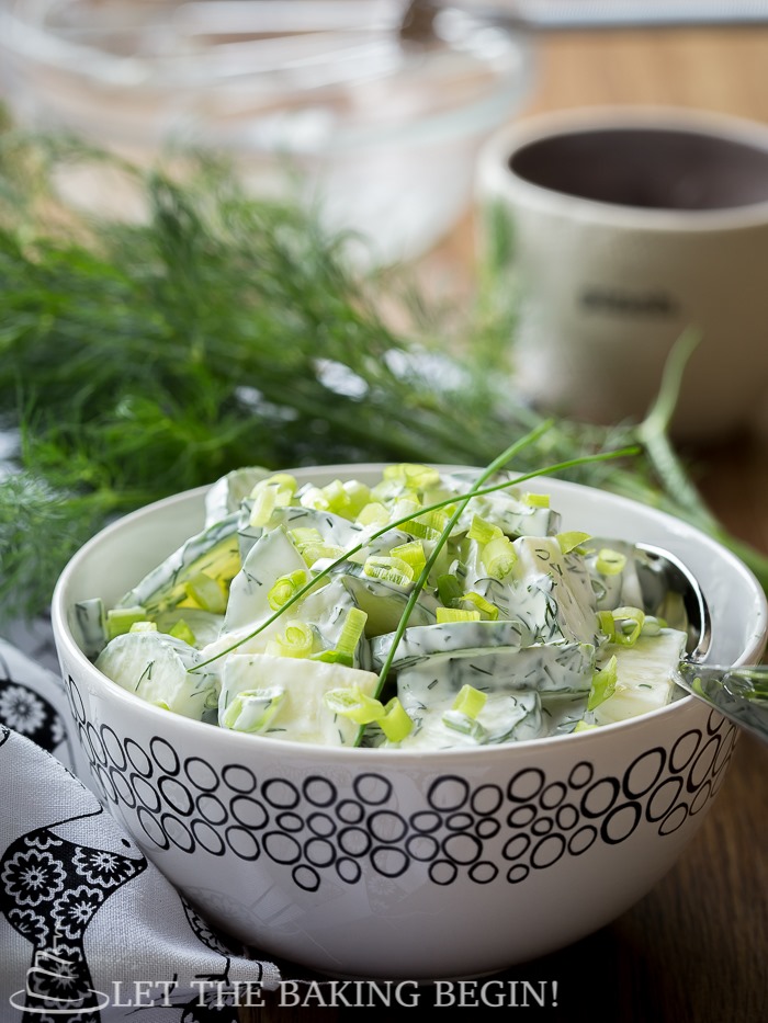 Side view of cucumber dill salad topped with fresh greens in a white decorative bowl on a wooden table. 