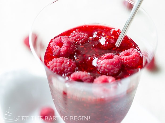 Homemade raspberry jam in a glass cup with a spoon topped with fresh raspberries. 