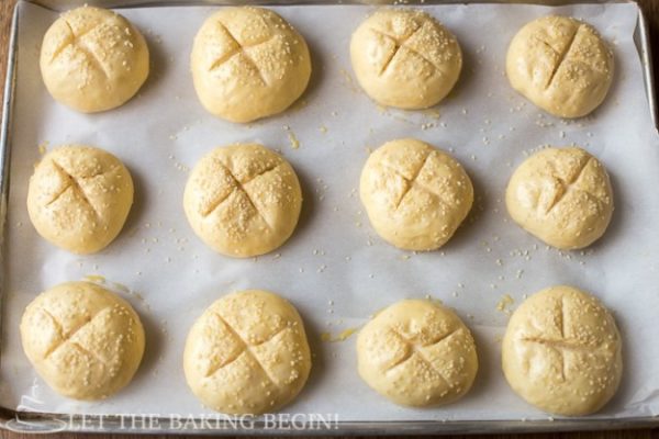  Top view of buns topped with sesame seeds and criss-cross incision on a parchment lined baking sheet.