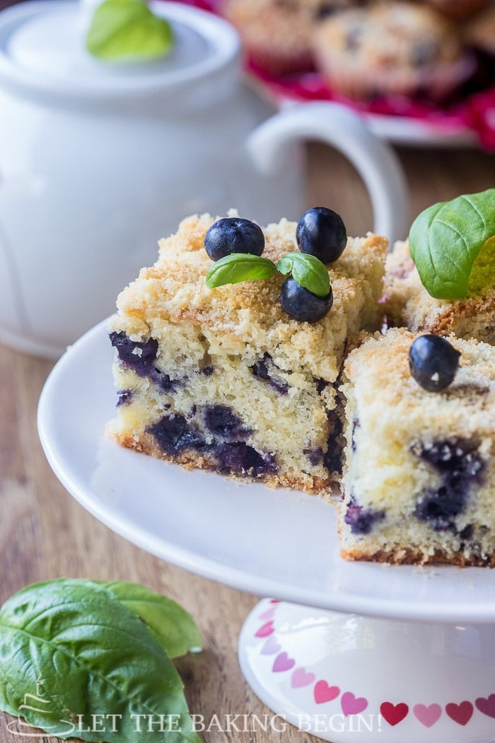  Blueberry cake topped with streusel toppings, blueberries, and mint on a white tray. 