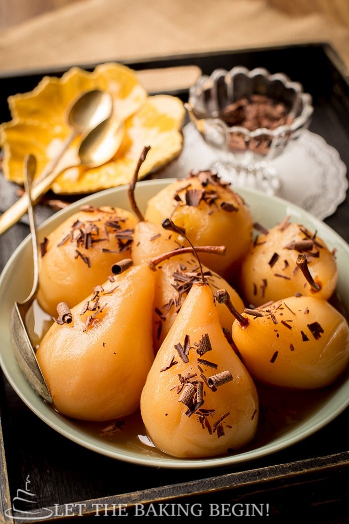 Poached pears topped with chocolate curls in a light green bowl in a wooden tray.