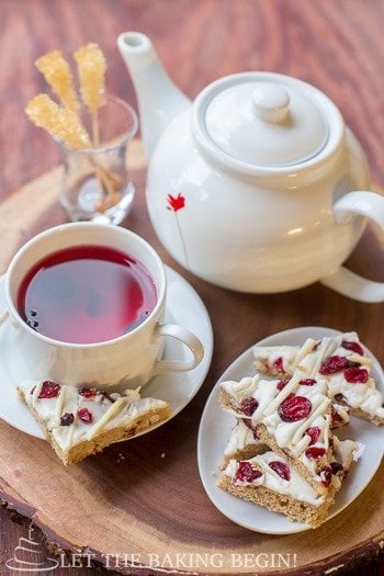 Cranberry bliss bar on a white plate with a cup of tea in a white mug next to a tea kettle, and another plate of cranberry bliss bars.