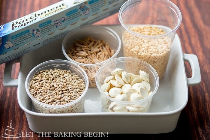 Parchment paper, white chocolate, sunflower seeds, almond sliver, and rice krispies in a white baking dish. 
