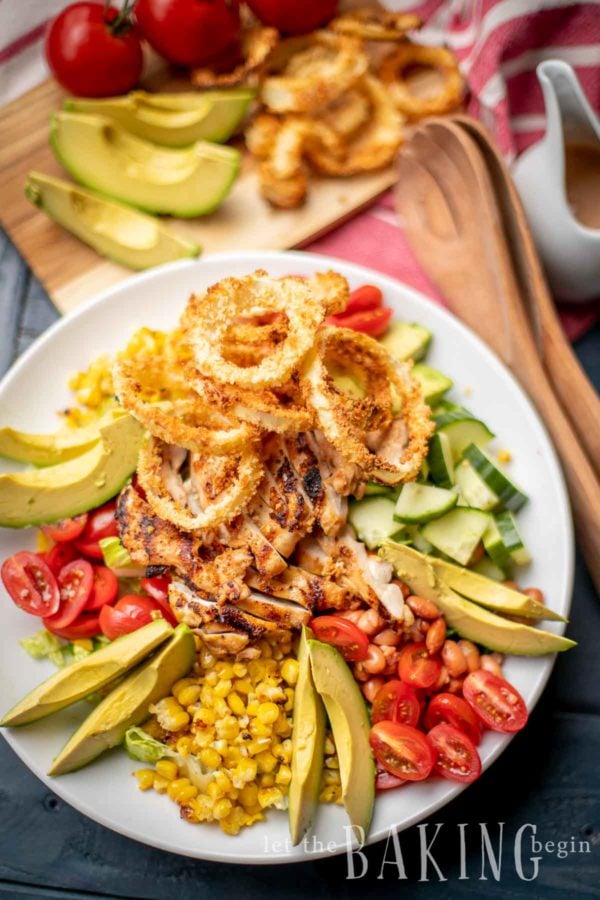 BBQ Chicken salad on a plate with avocado, tomato, corn, cucumber and fried onion rings.