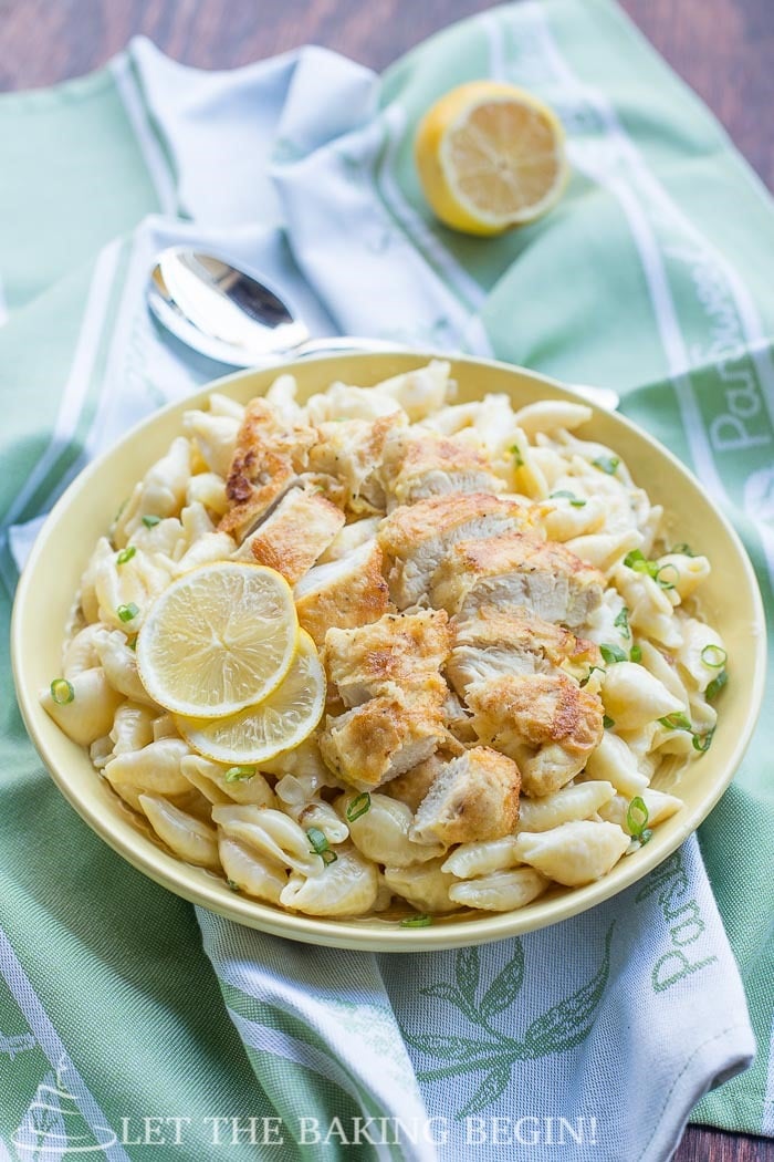 Cooked pasta topped with chicken tenders and fresh lemons in a yellow decorative bowl on a green napkin.