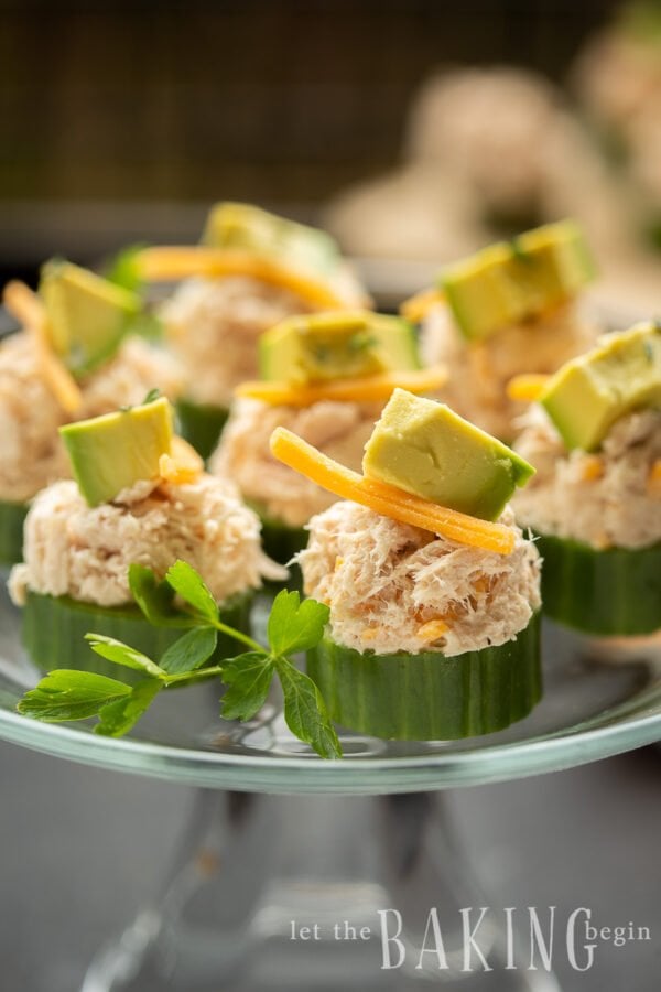 Seven cucumber tuna bites topped with cheese and avocado on a glass platter topped with fresh greens.