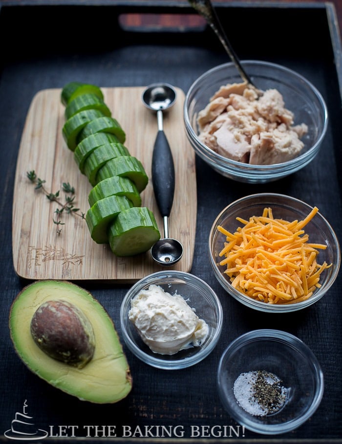 Sliced cucumbers, bowl of tuna, bow of cheese, a bowl of salt and pepper, bowl of mayonnaise, chopped greens, melon scooper and avocado displayed in a wooden tray.