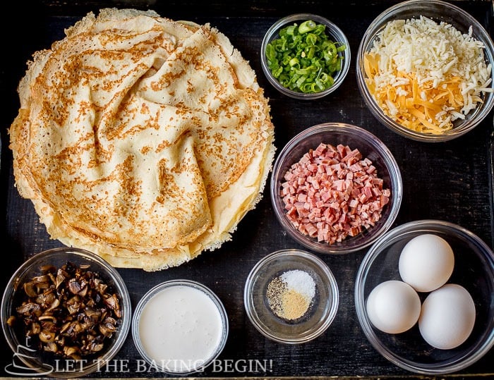 Cooked crepes, chopped mushrooms, diced ham/polish sausage, cheese, chopped chives or scallions, eggs, cream or milk, salt, pepper, garlic powder, and chopped parsley on a black wooden tray.