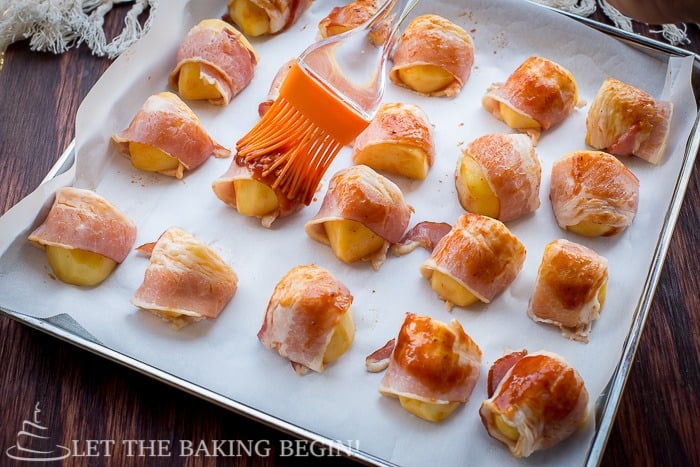 Bacon wrapped potatoes on a baking sheet with a pastry brush. 