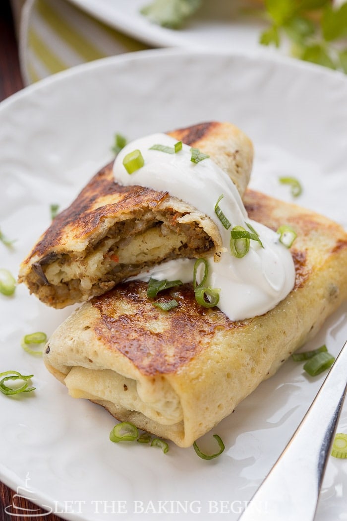 Two crepes topped with fresh greens and a dollop of sour cream on a white decorative plate.
