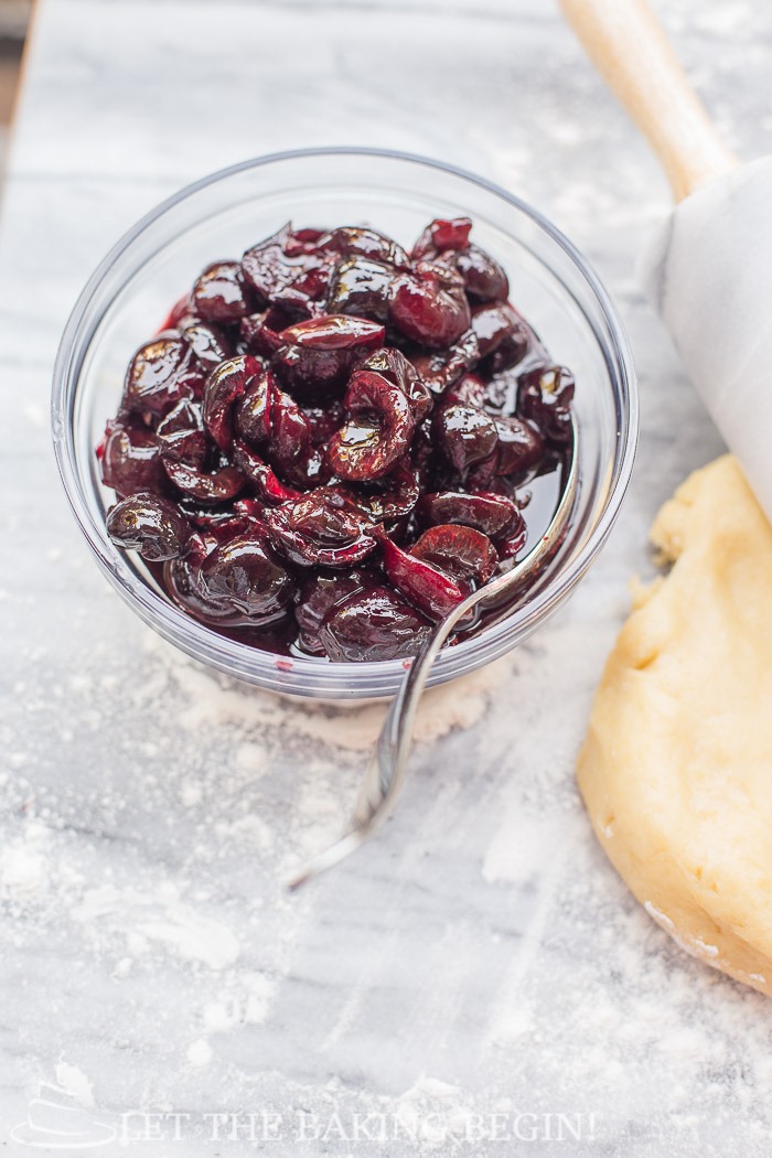Glass bowl of cherries with a spoon, next to homemade galette dough and rolling pin.