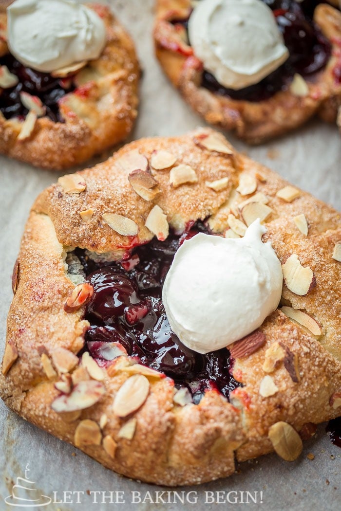 Cherry galettes with whipped cream topped with fresh almonds on parchment paper.