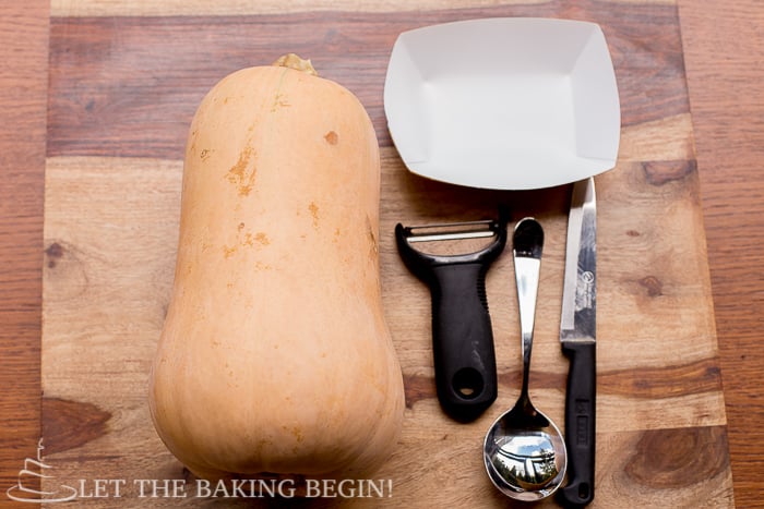 Butternut squash on a cutting board with a knife, spoon, and peeler. 