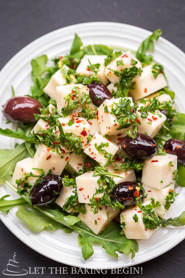 Marinated cheese appetizer on a plate with olives and topped with fresh chopped greens.