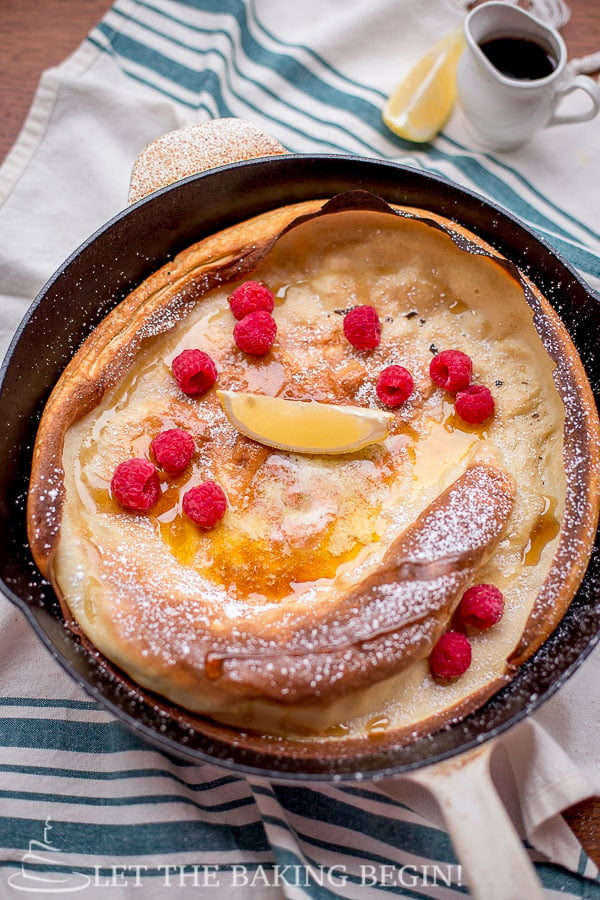 Shake and Bake German Pancake in a skillet topped with raspberries, syrup, and powdered sugar. 