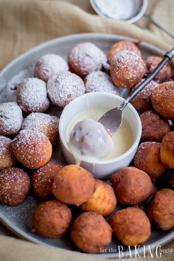 Doughnuts, half topped with powdered sugar and half plain, with icing in a bowl in the middle of a grey plate surrounded by doughnuts.