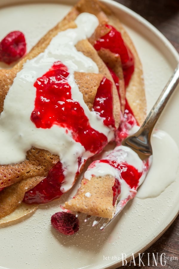 Crepes topped with raspberry and strawberry jam and sweetened sour cream with fresh raspberries on a white plate.