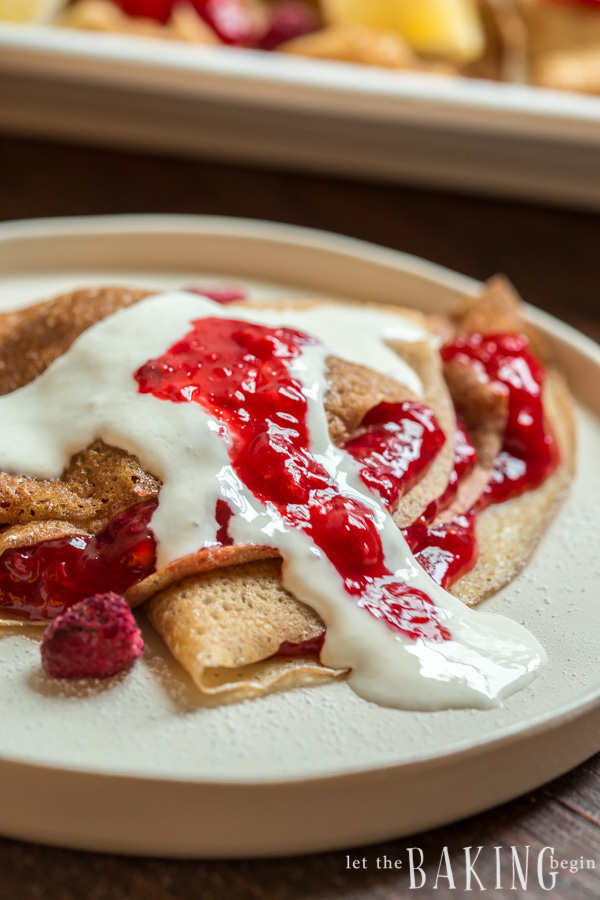 Crepes topped with strawberry and raspberry sauce and sweetened sour cream and fresh raspberries on a white plate.