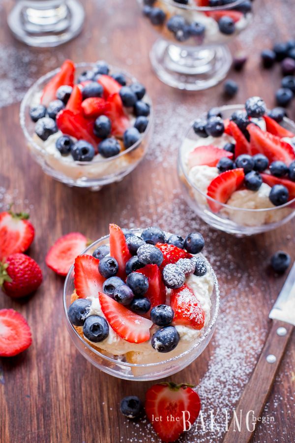 Cheesecake and berry trifles with blueberries and strawberries in glass cups topped with powdered sugar on a wooden board with cut strawberries and a knife