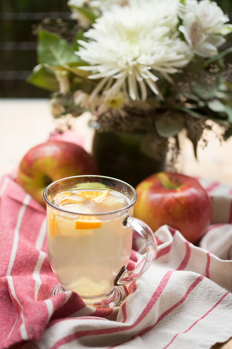 Apple drink with orange slices on a red and white towel with apples and a vase of flowers.