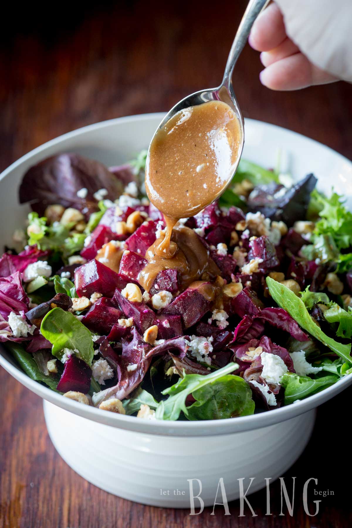 Salad topped with roasted hazelnuts, feta cheese, and vinaigrette being poured on the salad with a spoon in a white bowl. 