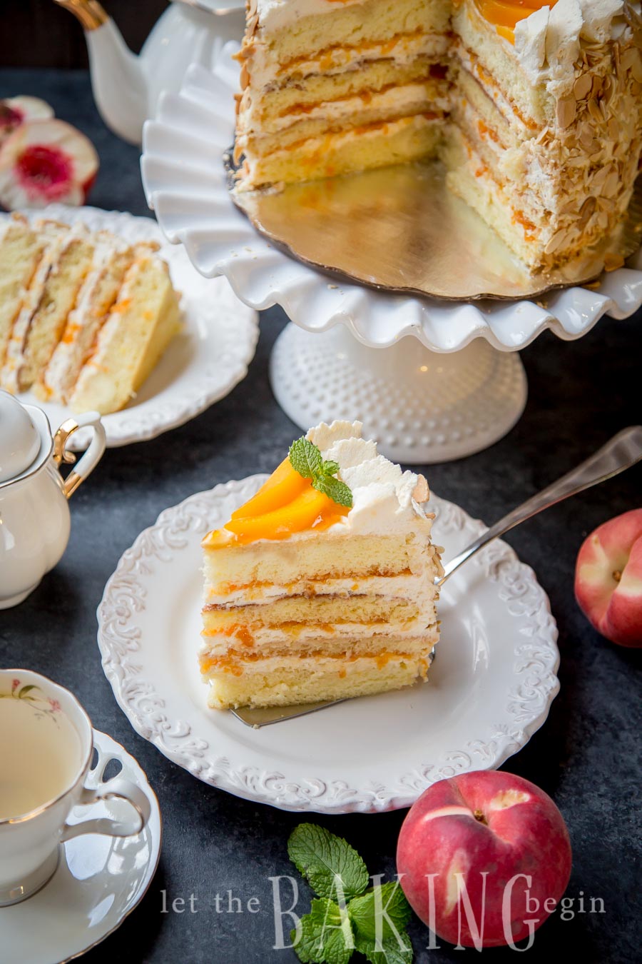 A slice of peached and cream cake on a plate next to a teacup and peaches. 