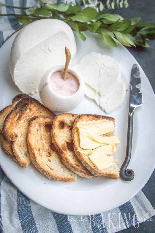 Block of farmers cheese, toasted bread with butter, butter knife and salt in a bowl on a white plate.