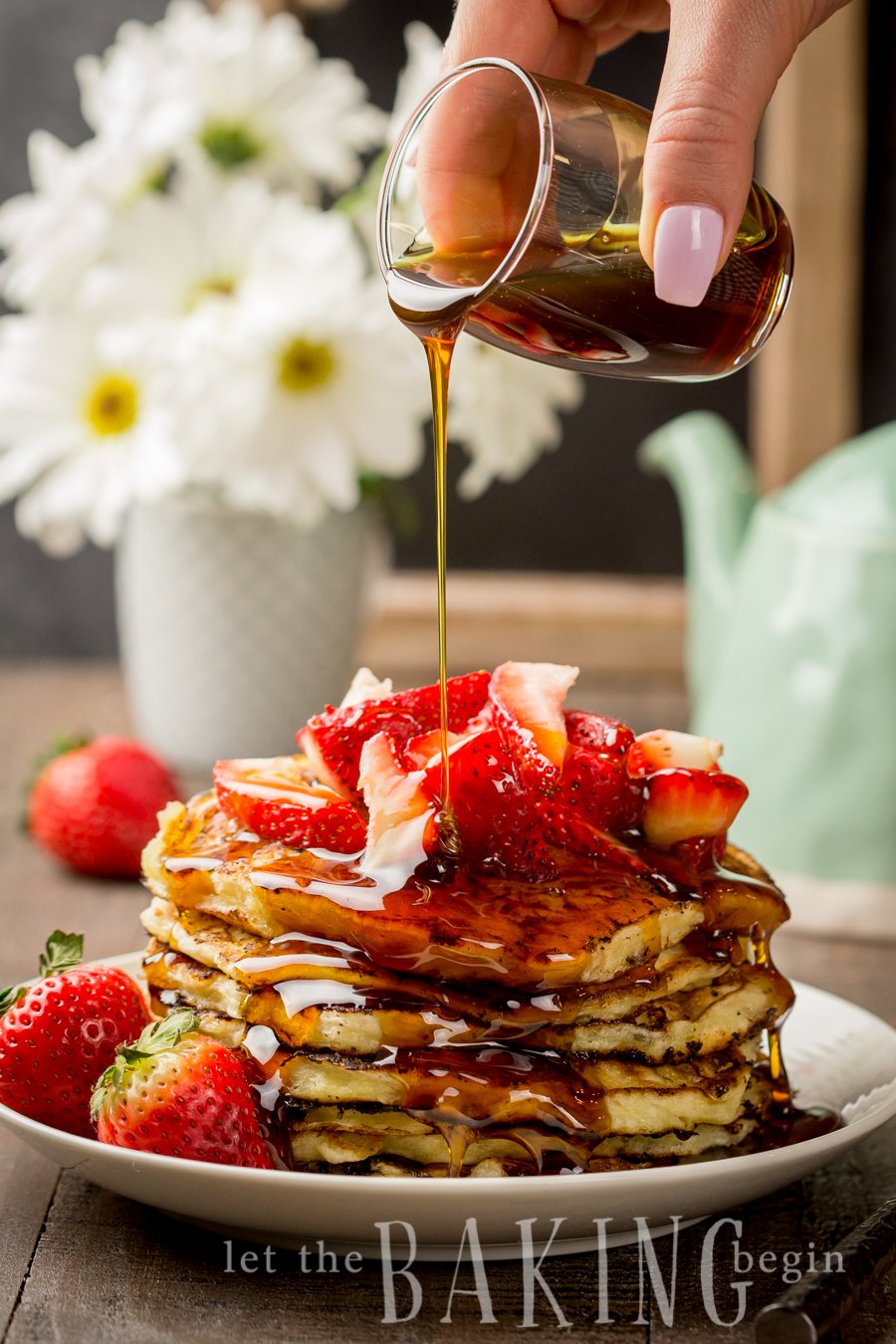 Maple syrup being poured onto a plate of cottage cheese pancakes topped with fresh strawberries. 