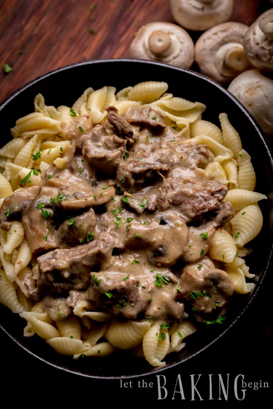 Beef stroganoff topped with fresh greens in a black bowl on a wooden table sided with mushrooms. 