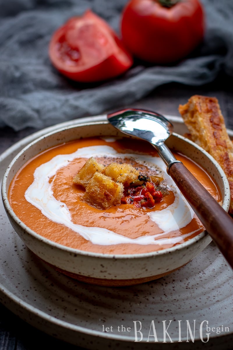 A bowl of roasted tomato soup topped with croutons and ground black pepper with a spoon. 