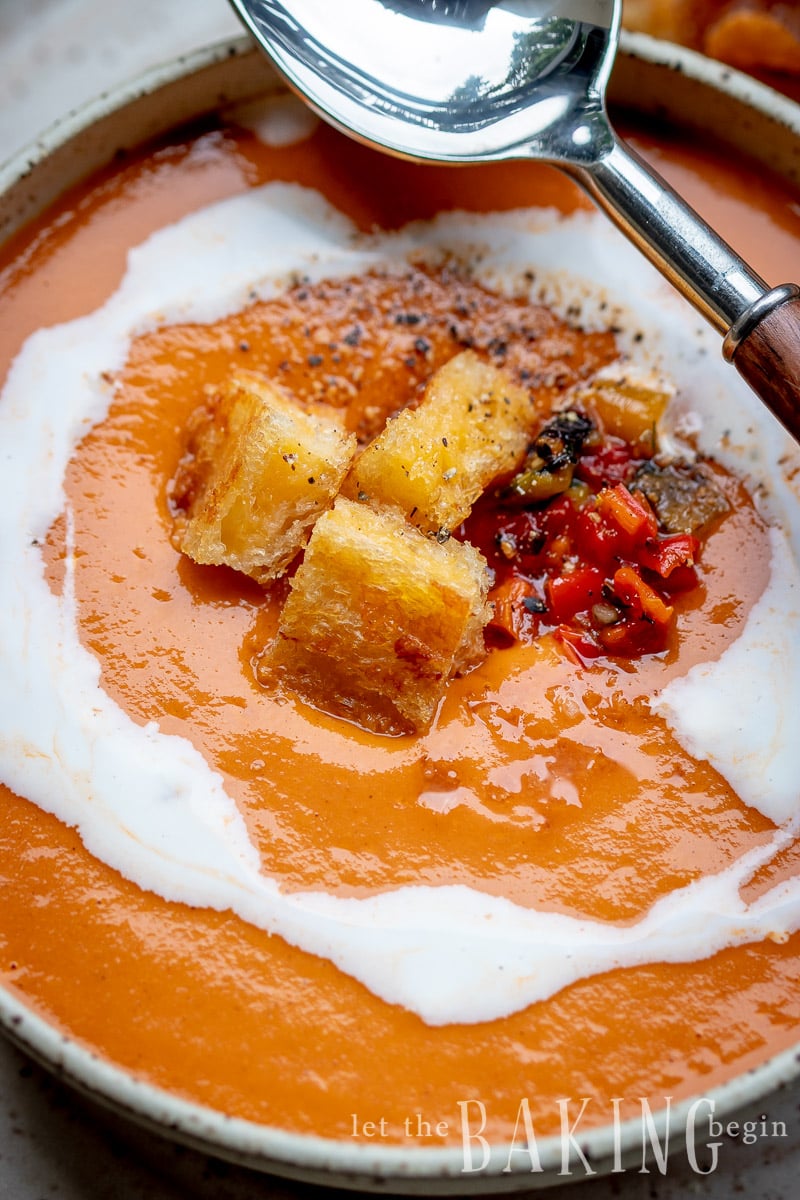 Creamy tomato soup topped with croutons and black pepper and red peppers with a metal spoon. 