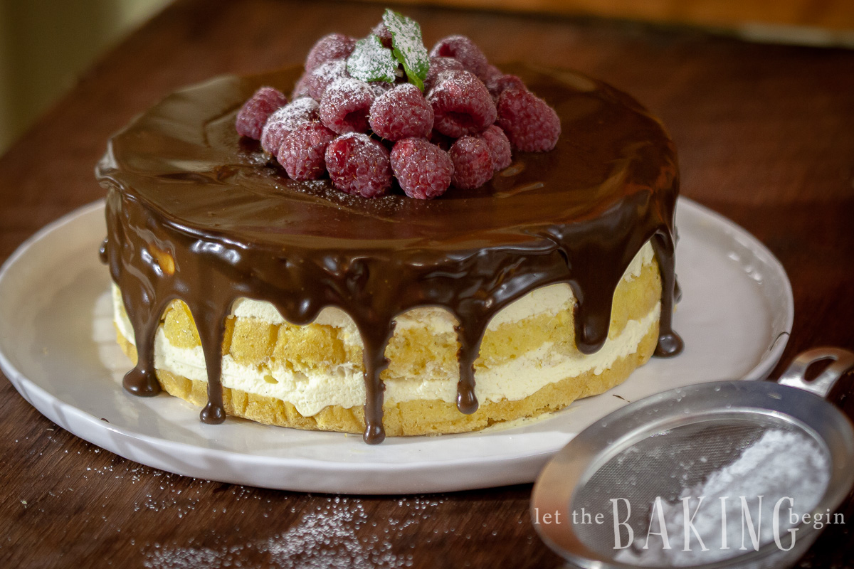 Boston cream pie recipe on a cake platter topped with chocolate ganache and fresh raspberries sprinkled in powdered sugar. 