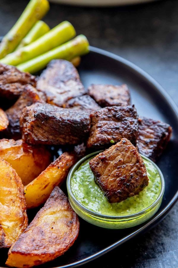 Steak bites with Asparagus, Roasted Potatoes on a plate, with one steak bite dipped in chimichurri sauce.